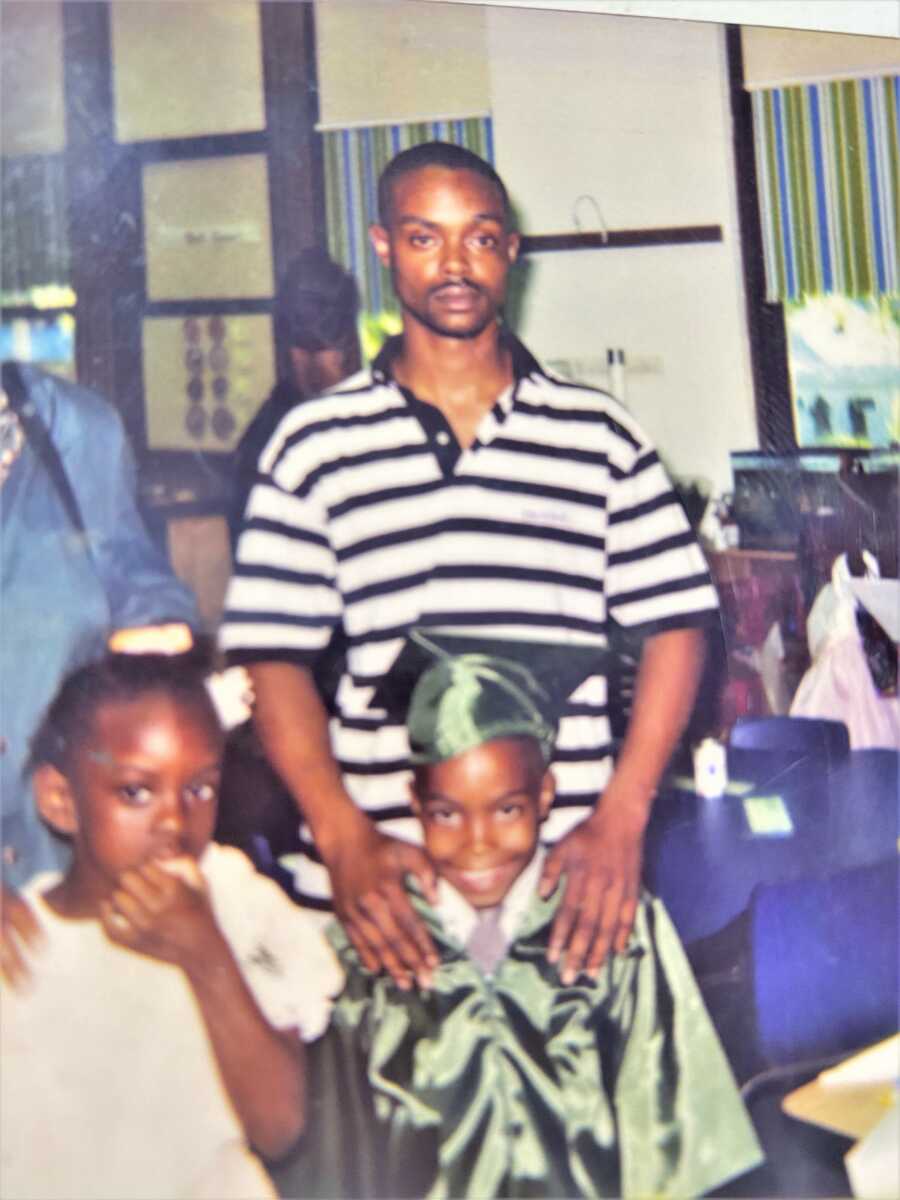 Black father standing behind his young son who is wearing a graduation gown and cap next to his little sister 