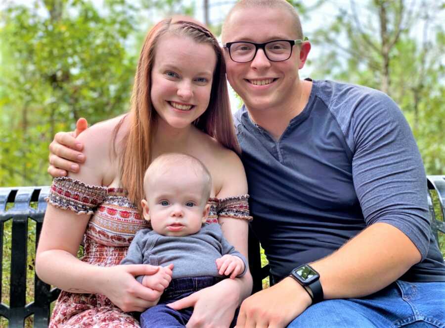 Dad and mom holding her baby son on her lap and smiling at the camera 