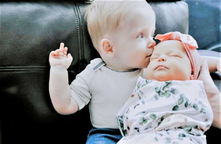 toddle boy holding his baby sister on his lap and giving her a kiss on the forehead