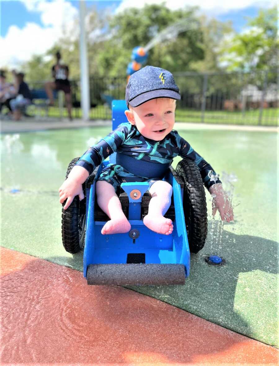 toddle boy with Spina Bifida sitting on special wheelchair playing with the water of a fountain 