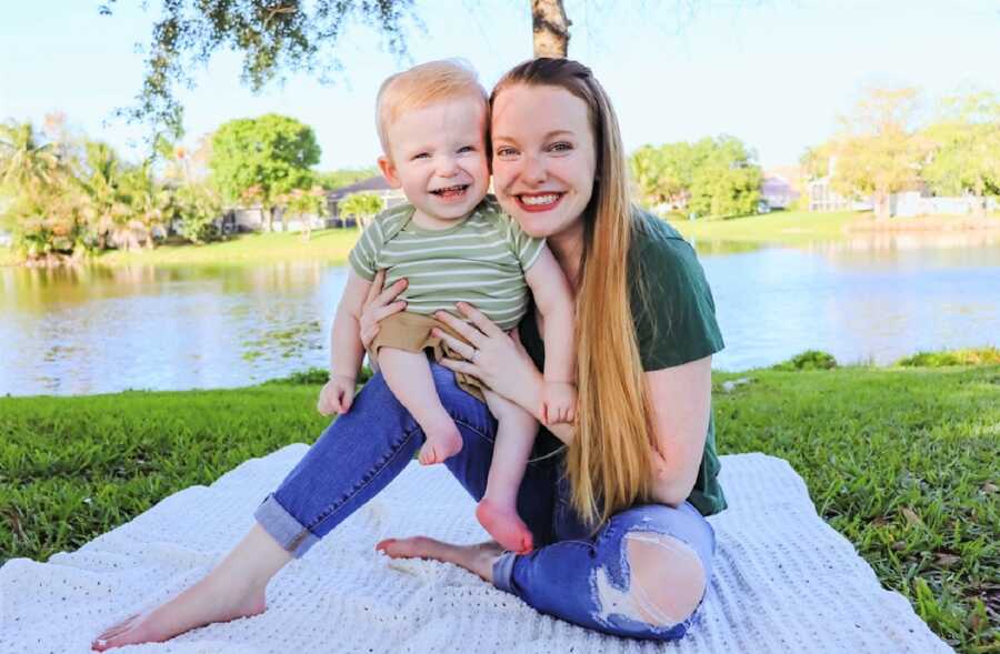 mom and toddler son smiling while sitting on a white blanket laying on top of the grass at a park 