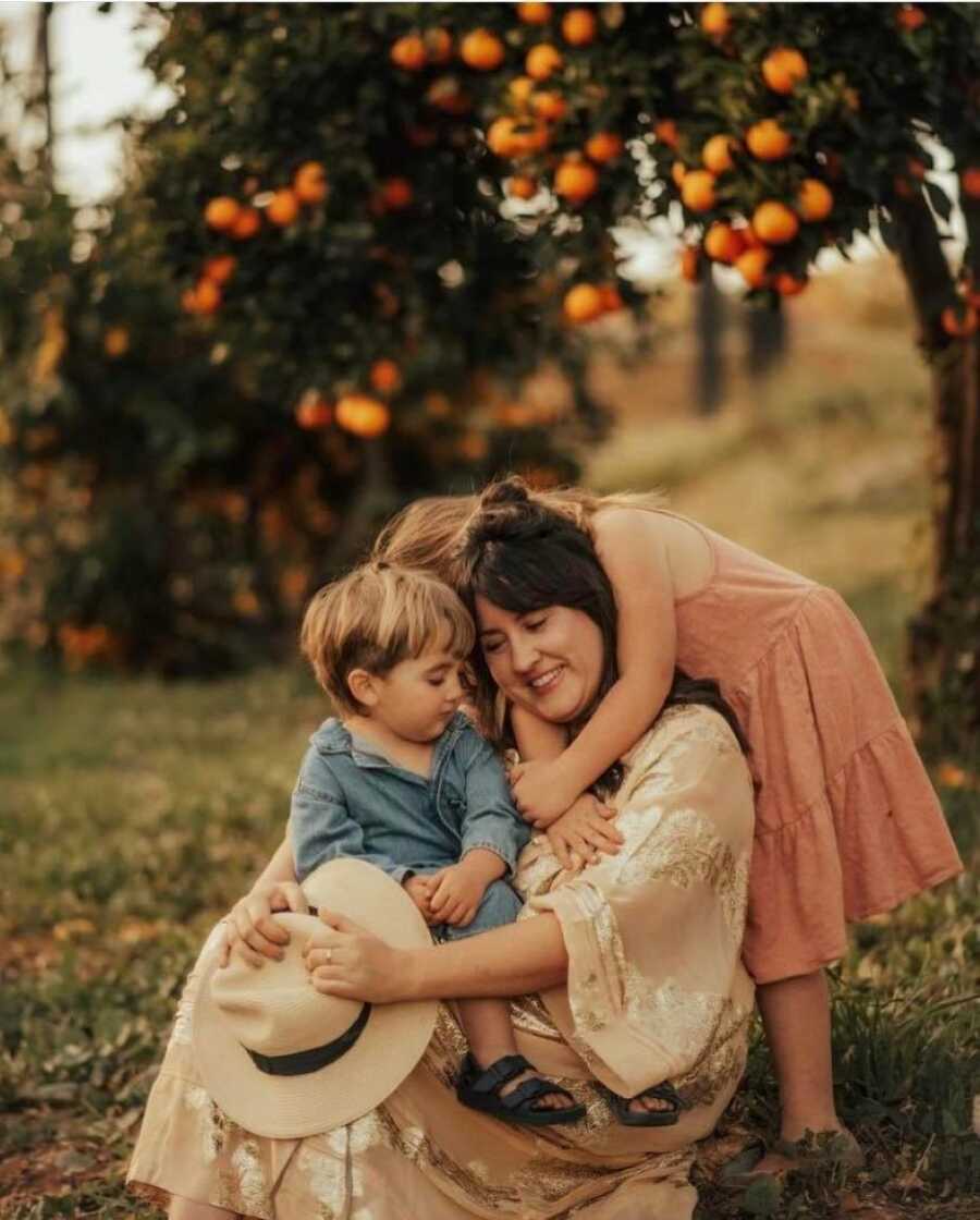 Mom hugs her two children while sitting on a farm with orange trees behind them