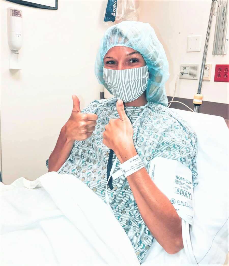 Woman dressed in patient apparel sitting at a hospital bed with both of her thumbs up 
