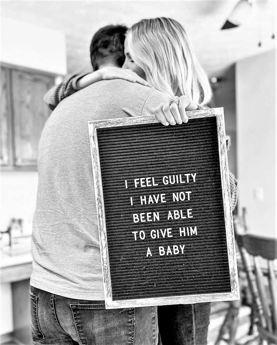 husband and wife hugging while wife holds a sign that says" I FEEL GUILTY I HAVE NOT BEEN ABLE TO GIVE HIM A BABY"