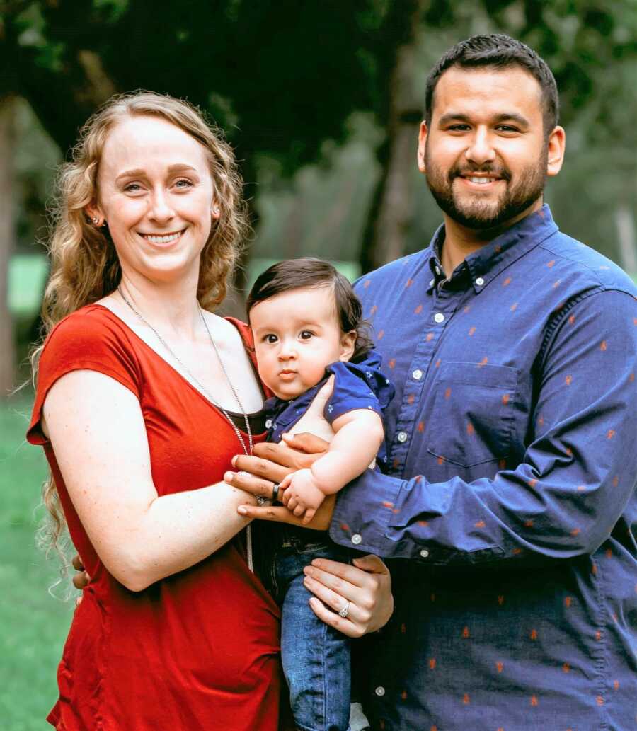 Professional portrait of mom and dad holding their baby son and smiling with trees in the background 