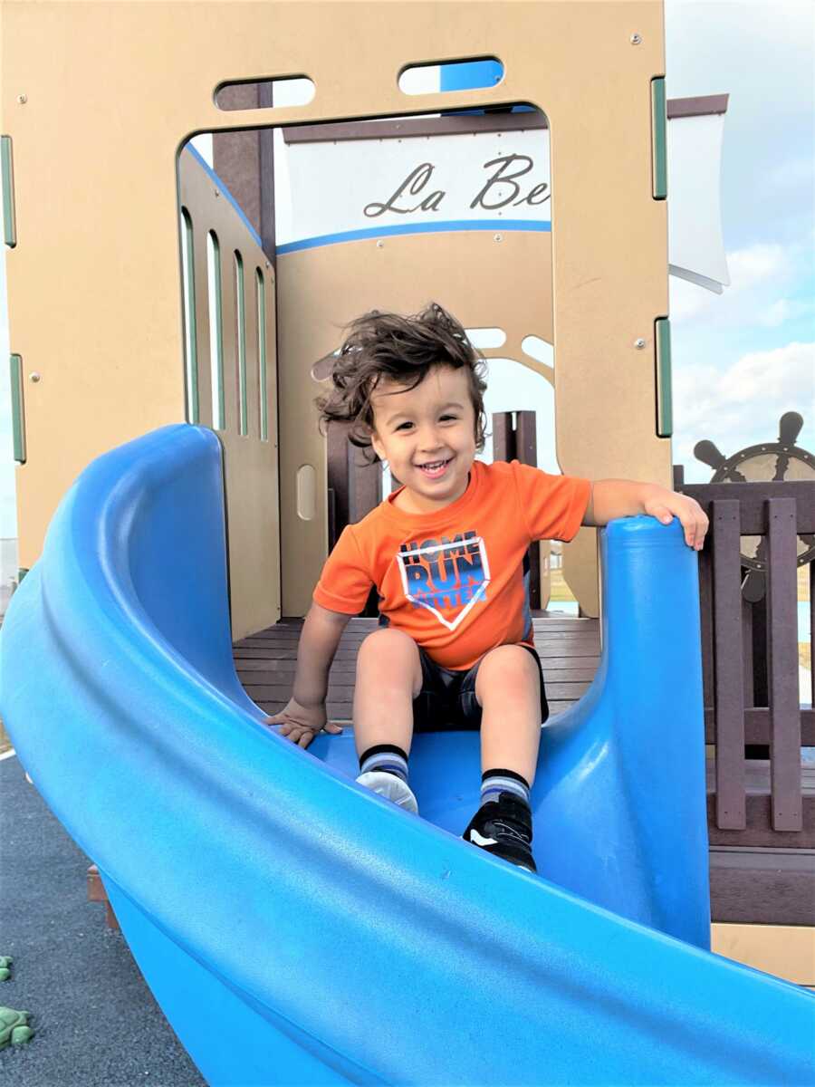 Baby boy sitting at the top of a blue slide at a playground 