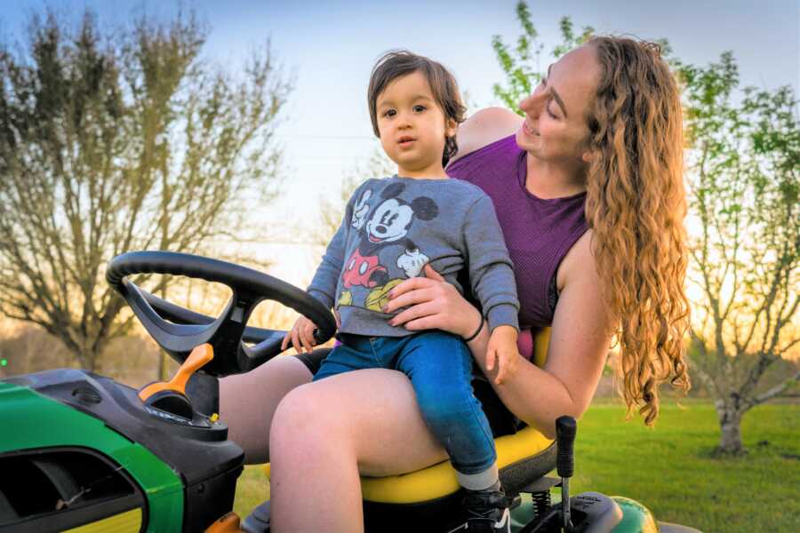Mom sitting on a green lawnmower with her toddler son on her lap