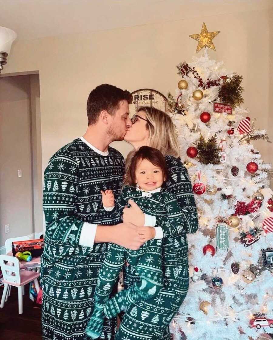 Couple share a kiss while holding their adoptive daughter in front of the Christmas tree in matching PJs