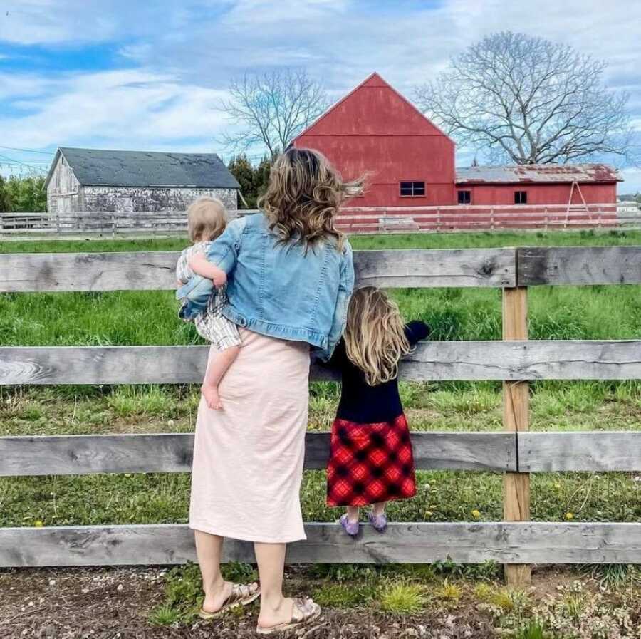 Mom and two daughters lean on a fence while looking out over their farm