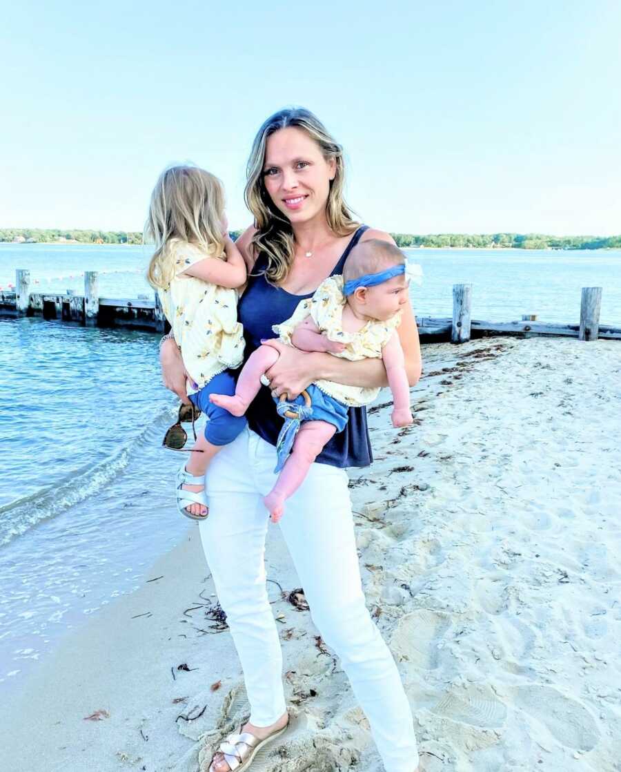 Mom holding her baby daughter and her toddler daughter in each arm while standing on the sandy shore of a lake 