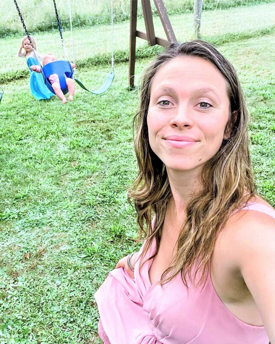 selfie of a mom with her two daughters in the background playing on a swing at a playground 