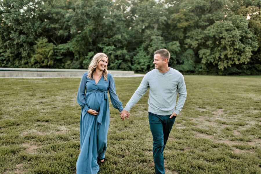 Pregnant woman wearing a long blue dress walking on a park holding her husband's hand 