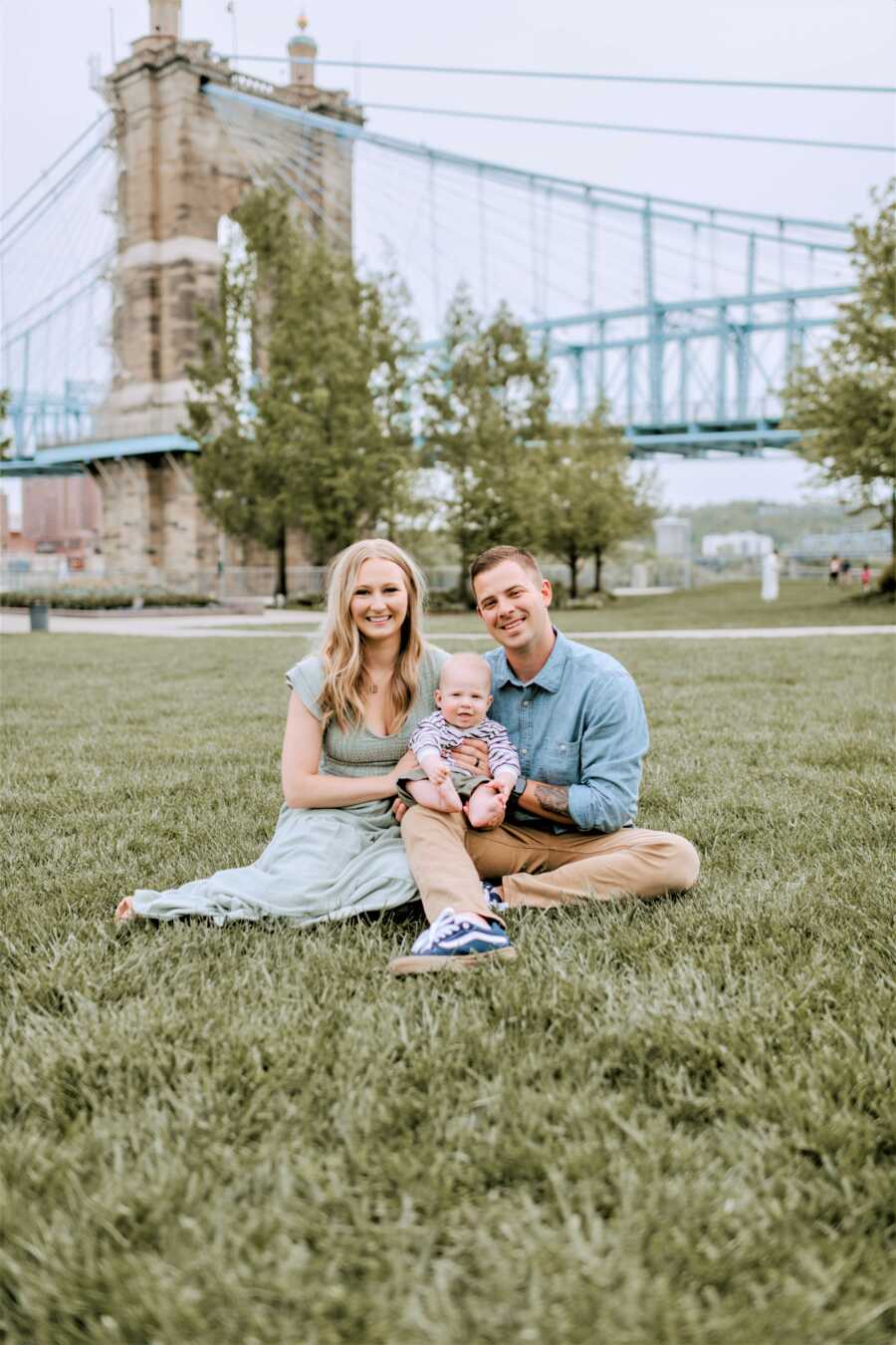 Outdoor family portrait of mom, dad, and baby son sitting on the grass with a bridge in the background 