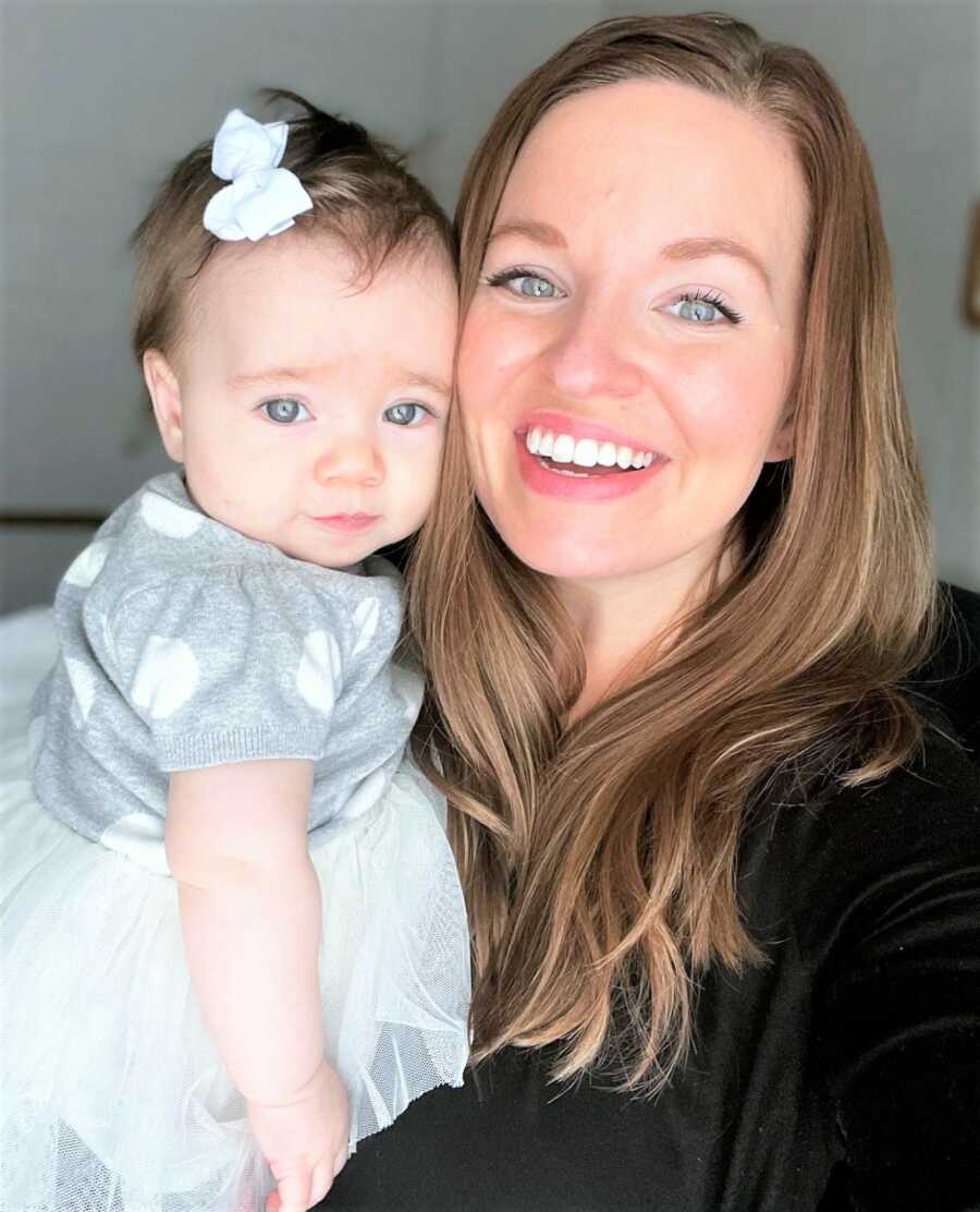 selfie of a mom smiling and holding her baby daughter who is wearing a white tutu and little bow on her head