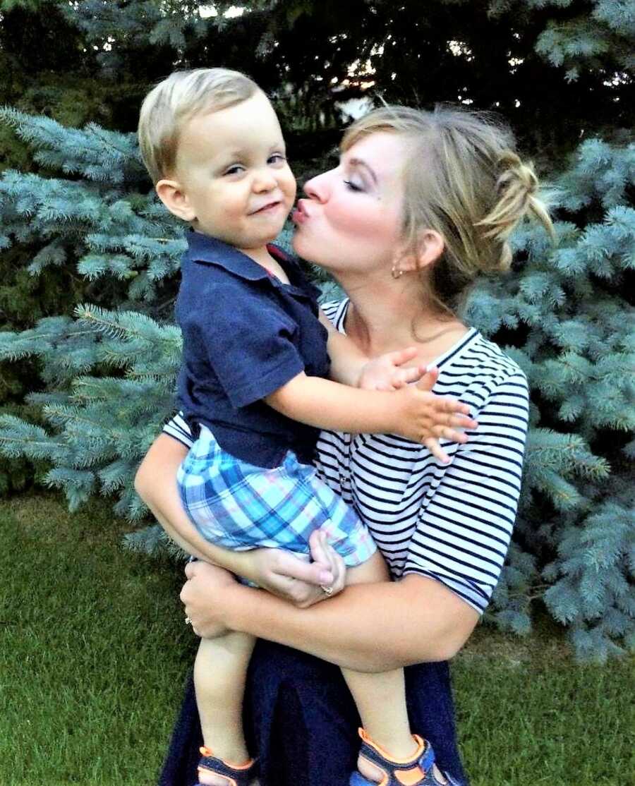Mom holding toddler son on her arms while giving him a kiss on the cheek 