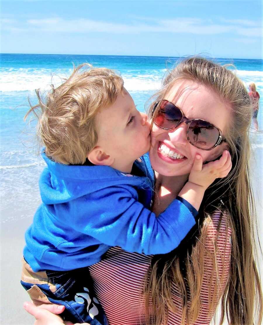 mom holding her toddler son in her arms while he gives him a kiss on the cheek at the beach 