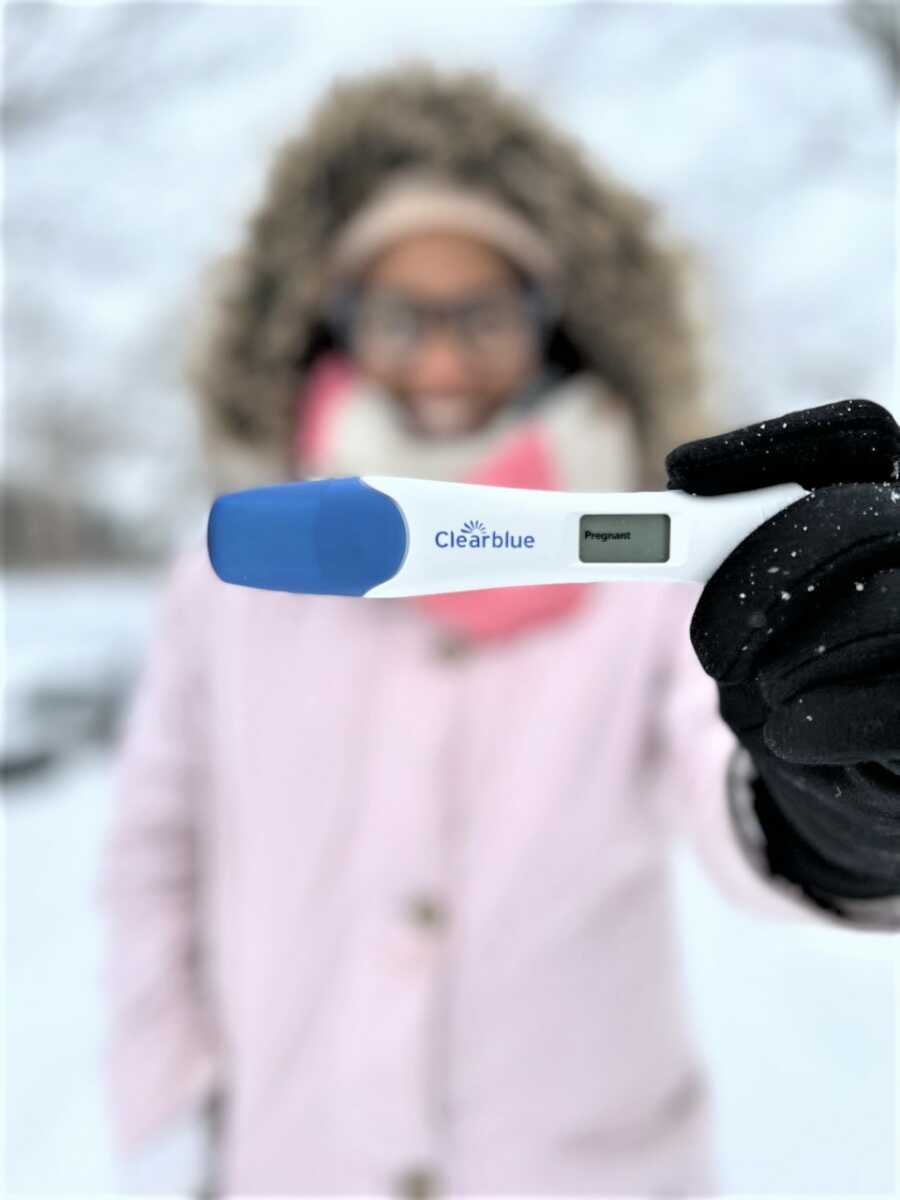 pregnant woman blurred in the back holding a digital pregnancy test that says 'pregnant'