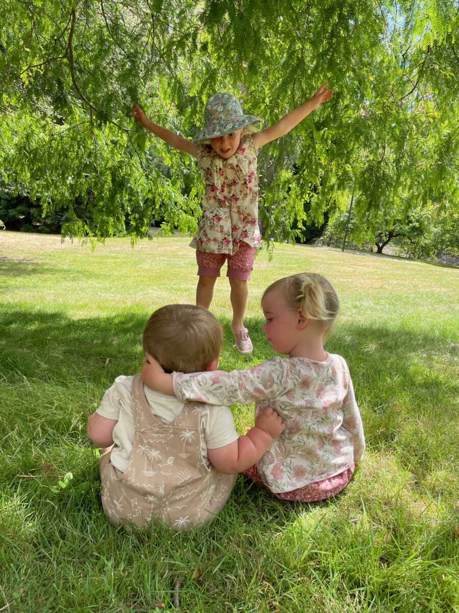 Sisters puts arm around younger brother as they sit outside watching older sister.