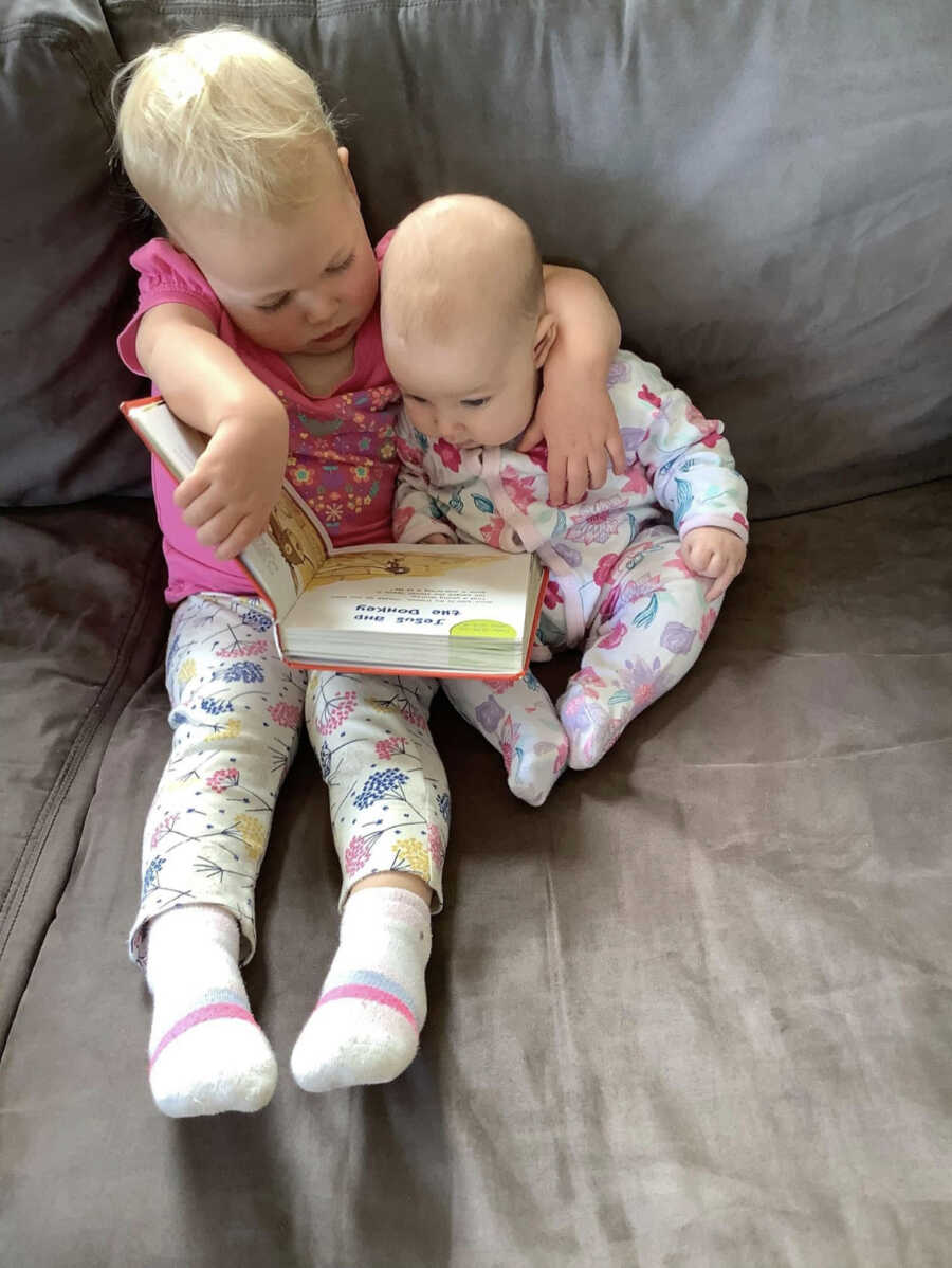 Big sibling sits with arm around baby on the couch and reads them a book.