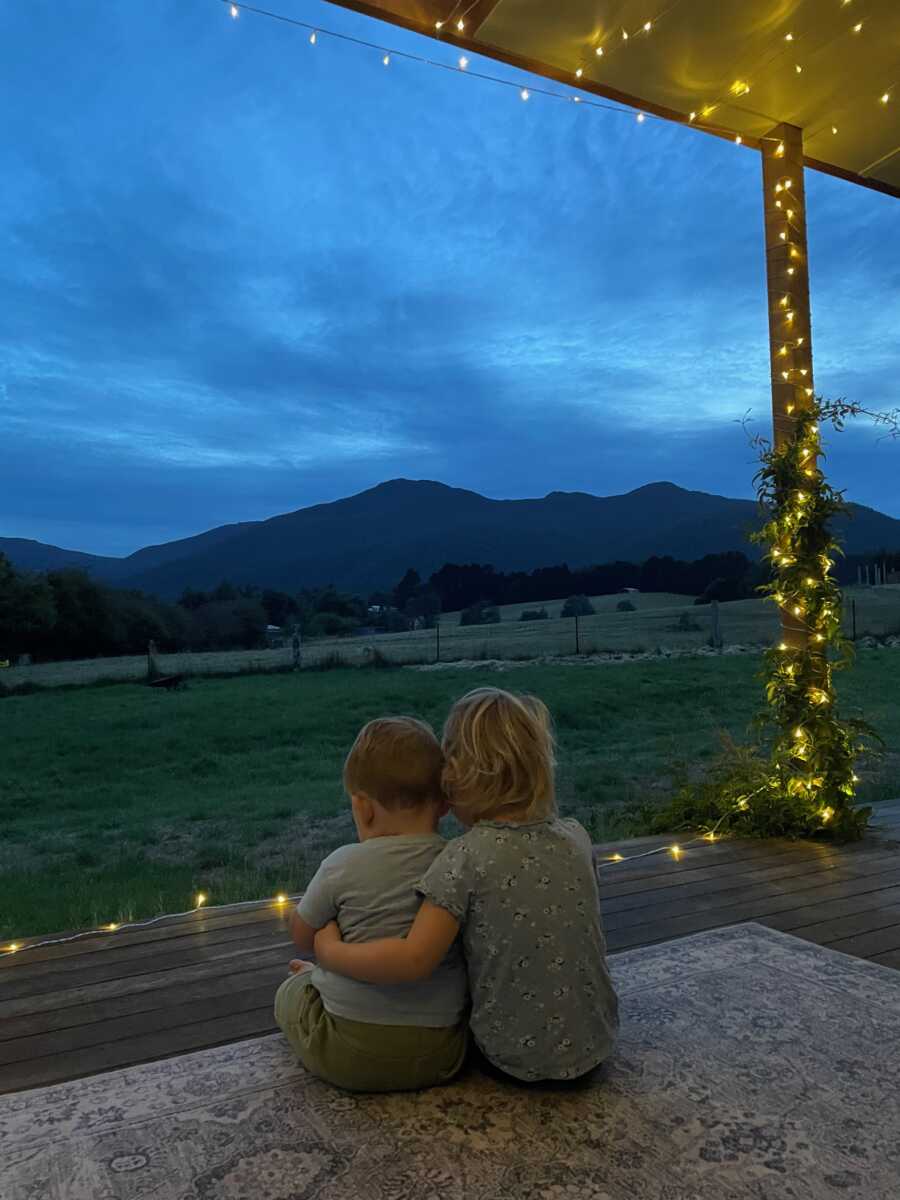 Foster siblings sit outside together at night, looking out over the farm land.