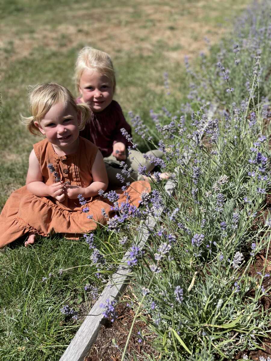 Sisters sit outside in the grass next to a flower bed and smile for a picture. 