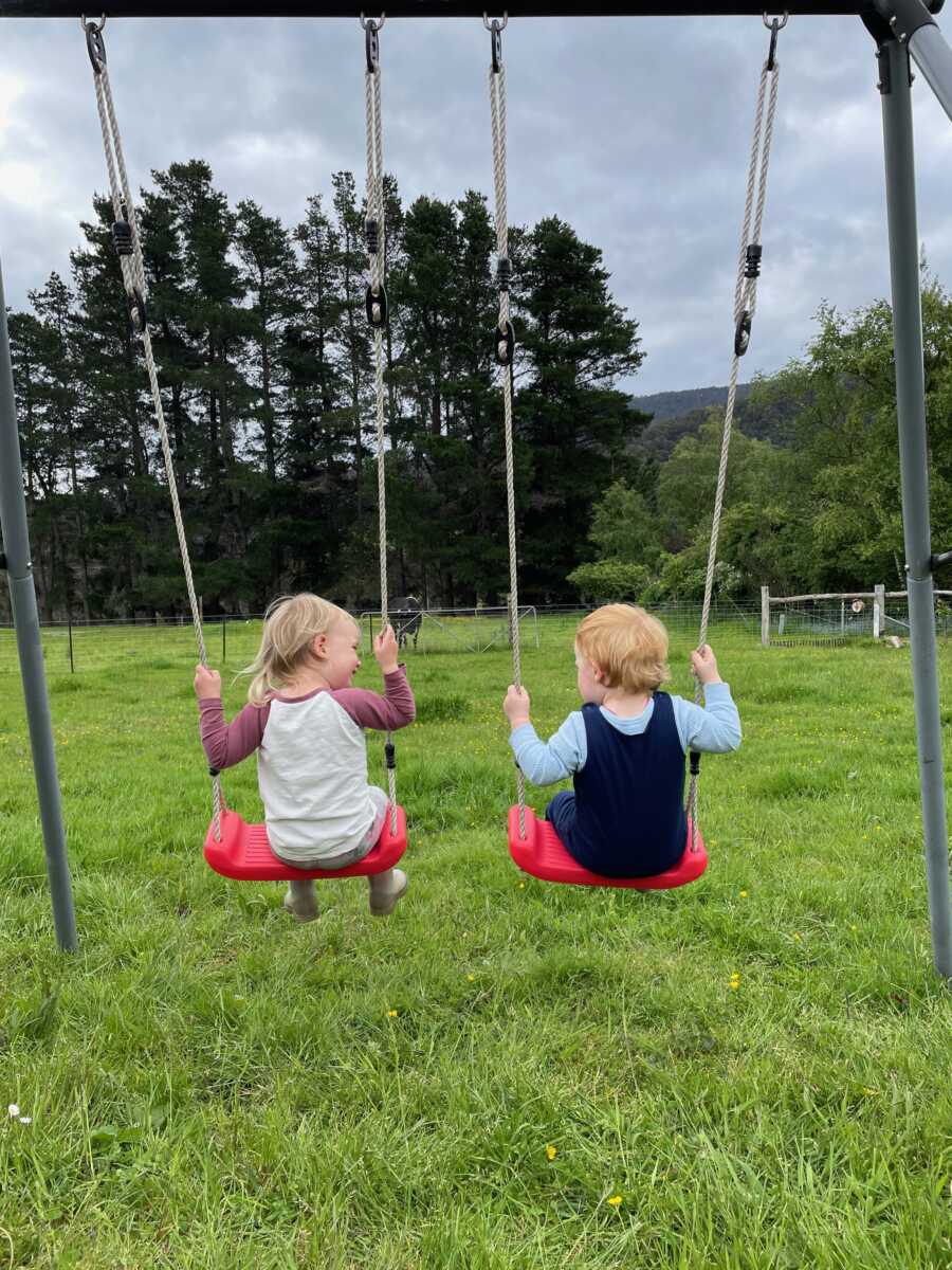 Young siblings swing next to each other on playground swings.