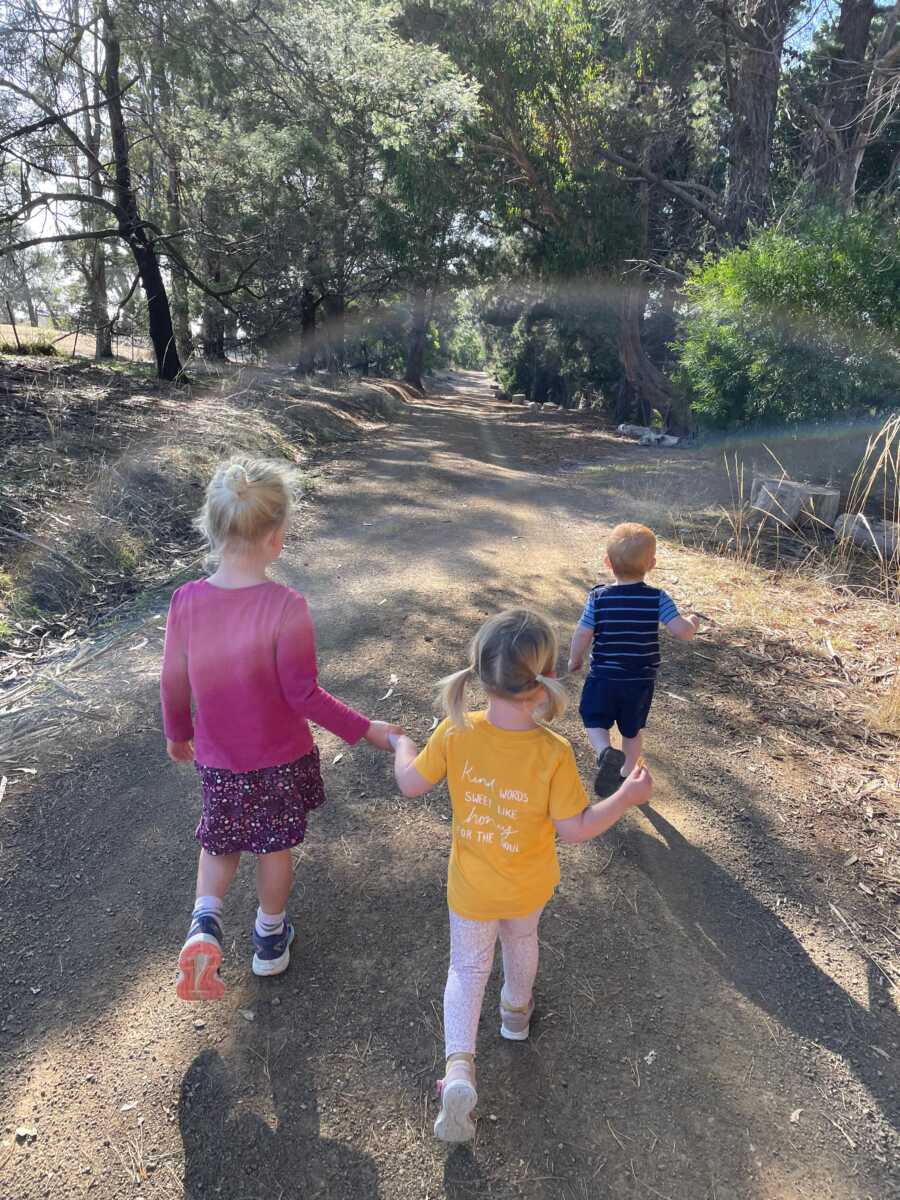 Young siblings hold hands as they hike down a wide forest path.