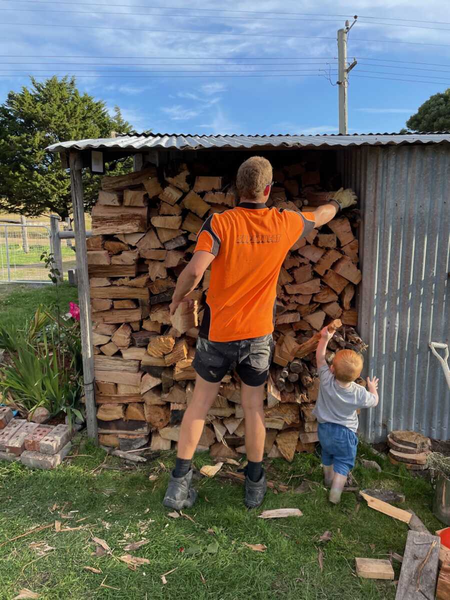 Husband stacks large firewood storage with toddler watching nearby.