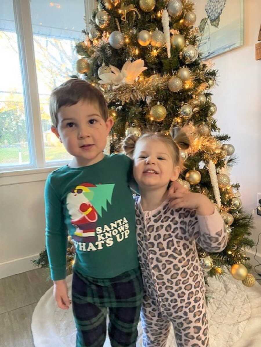 Brother puts arm around little sister as they stand in front of a Christmas tree decorated in gold.
