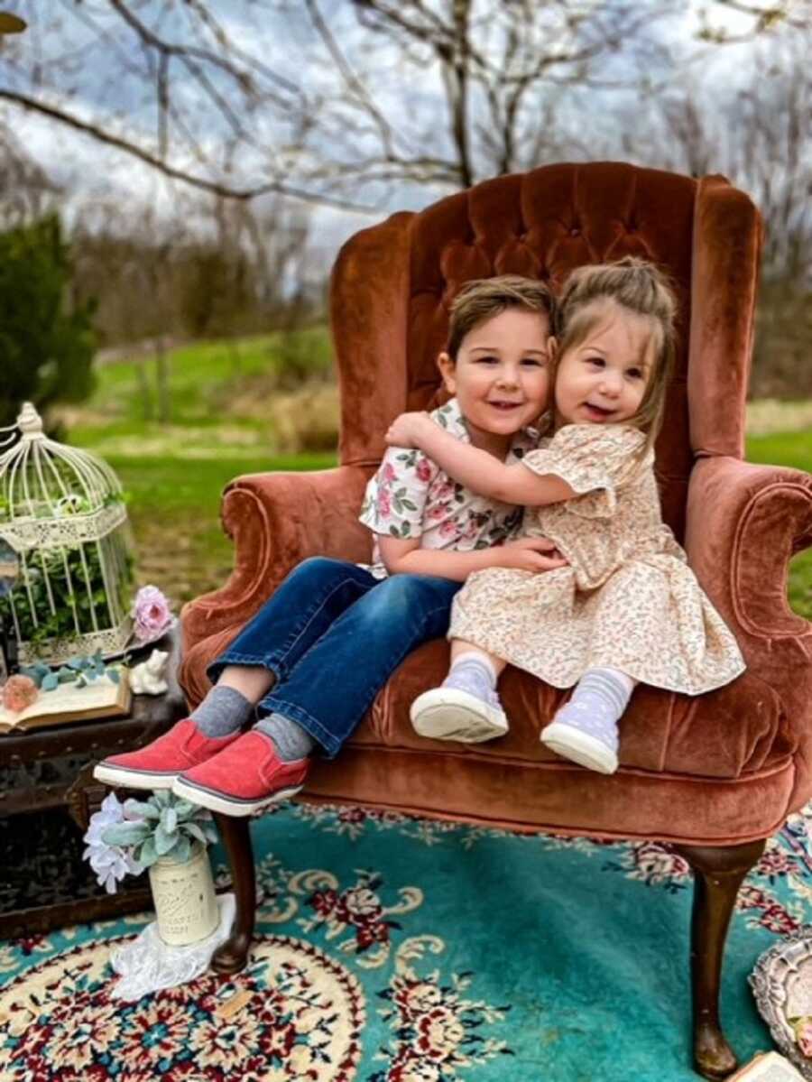 Adopted siblings sit in cushioned arm chair wrapping arms around each other.