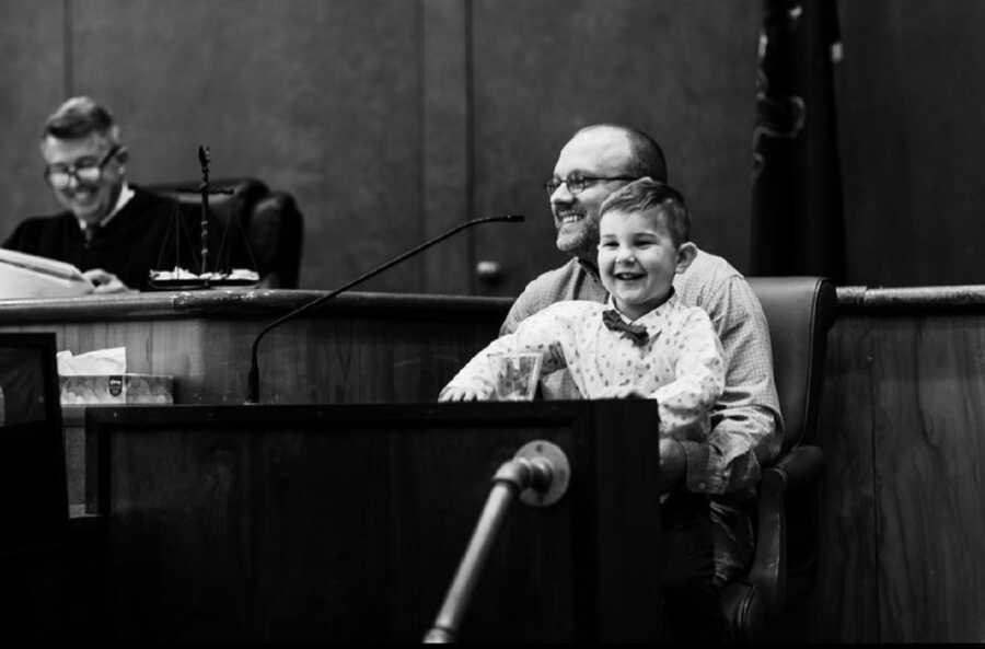 Young boy sits on dads lap in court room on adoption day.