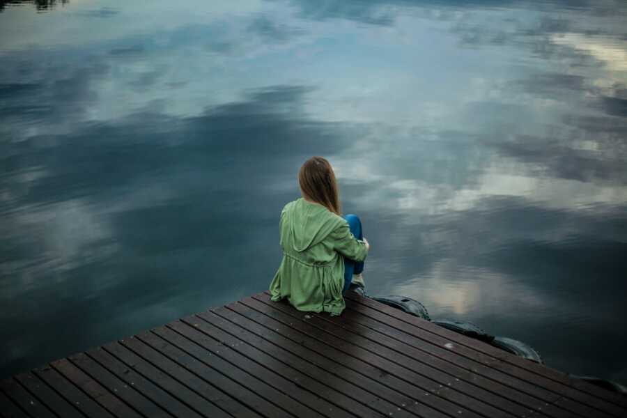A woman sits alone on a pier by the water wearing a green jacket