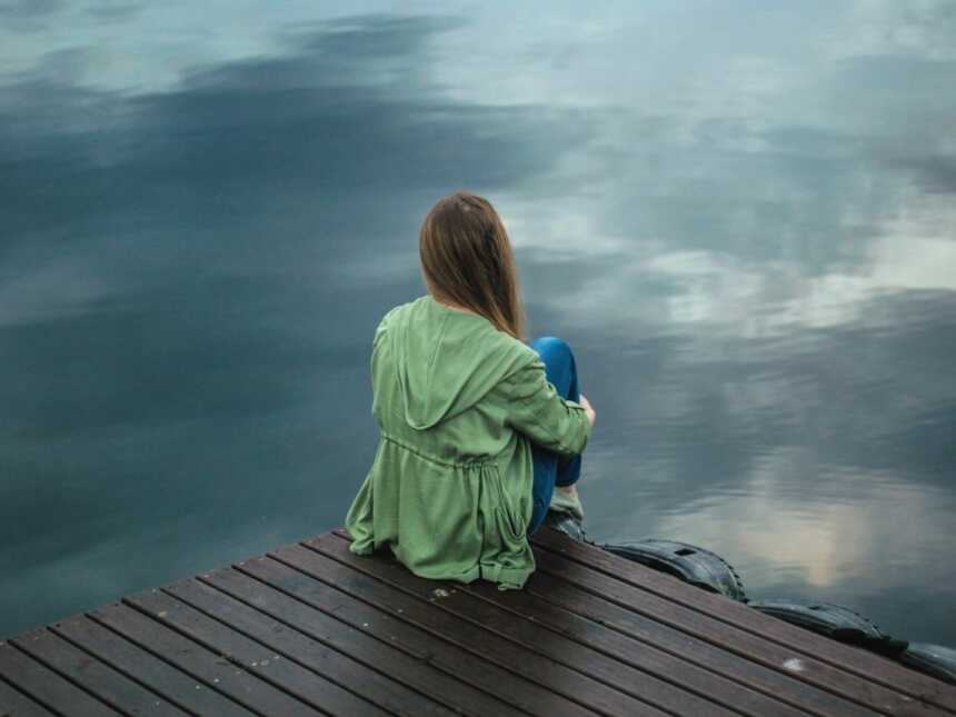 A woman sits alone on a pier by the water wearing a green jacket