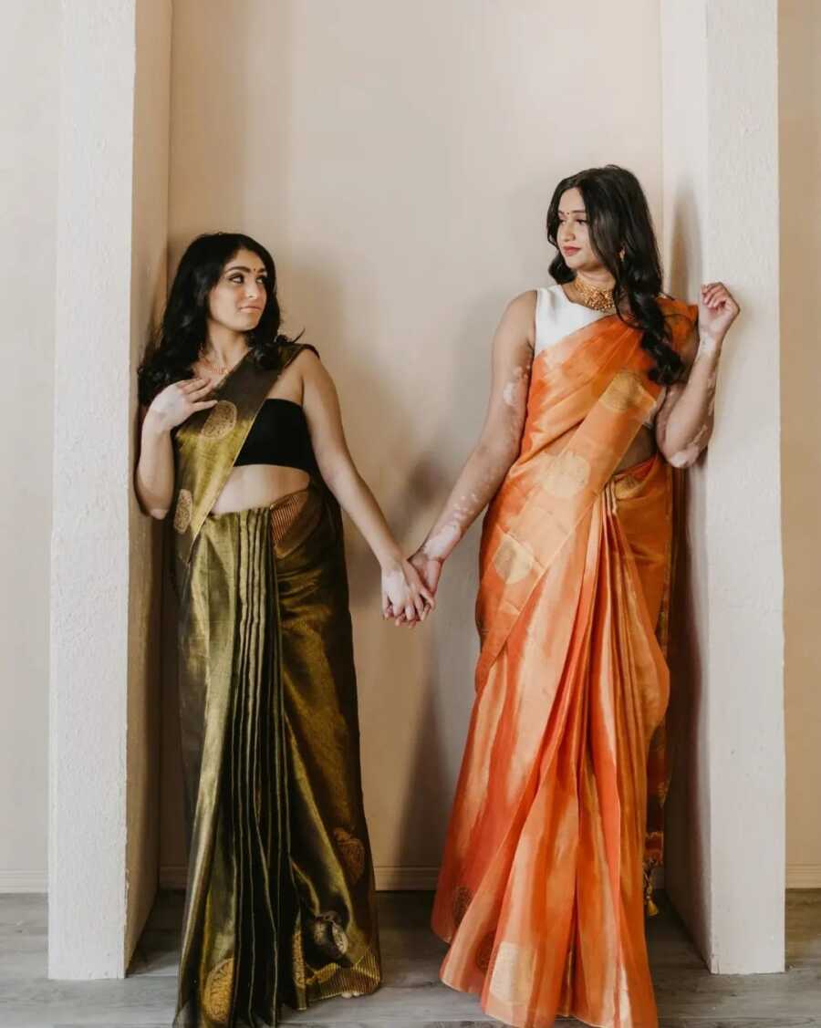 two woman with vitiligo stand against a wall holding hands