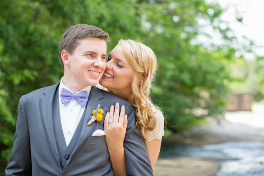 wife stands behind husband with her arms around him on their wedding day