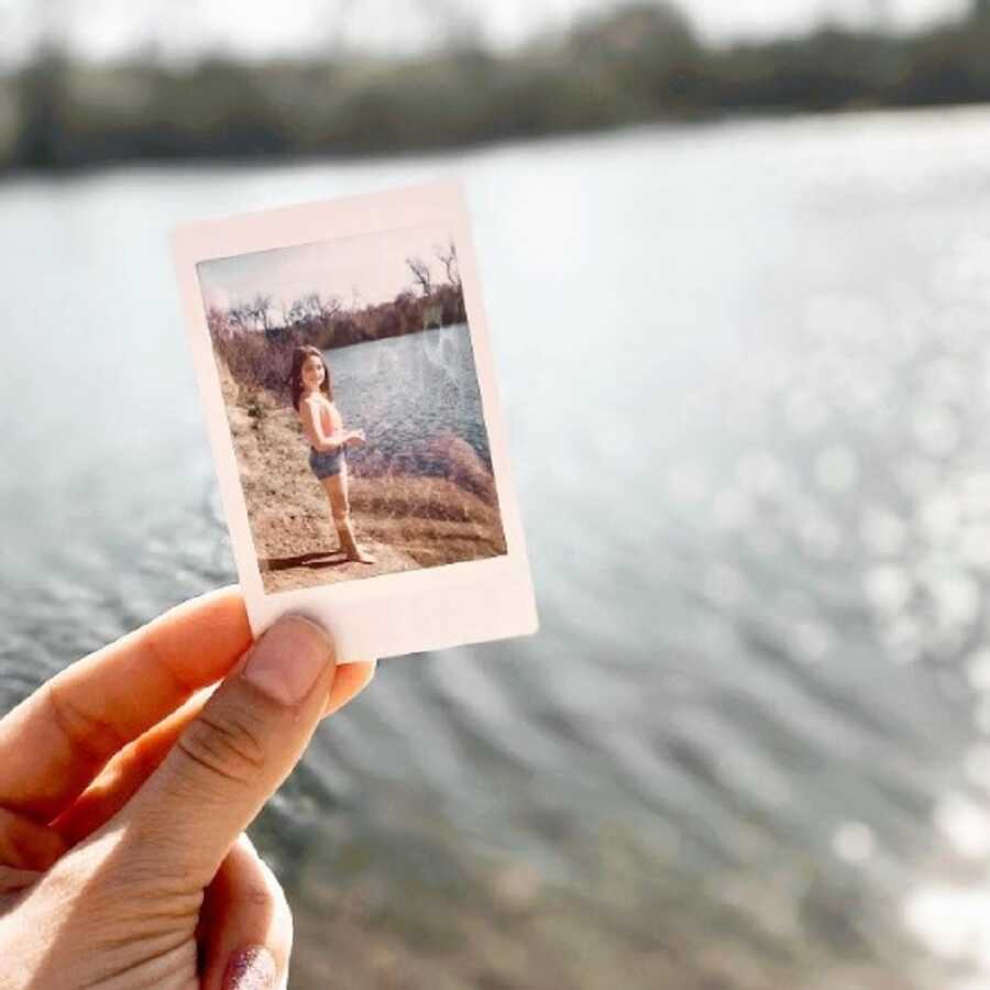 Woman holds picture of herself as a child against backdrop of a lake.