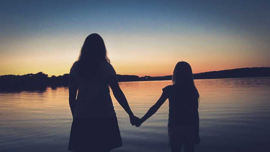 mother and daughter standing hand in hand facing the water