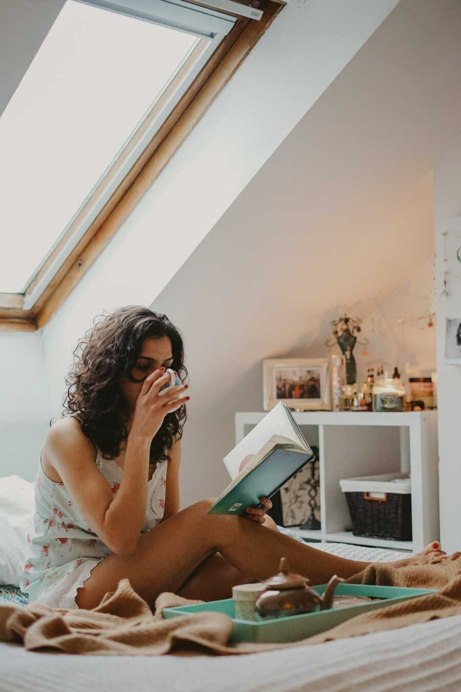 woman sits curled up in bed with a cup of coffee and book