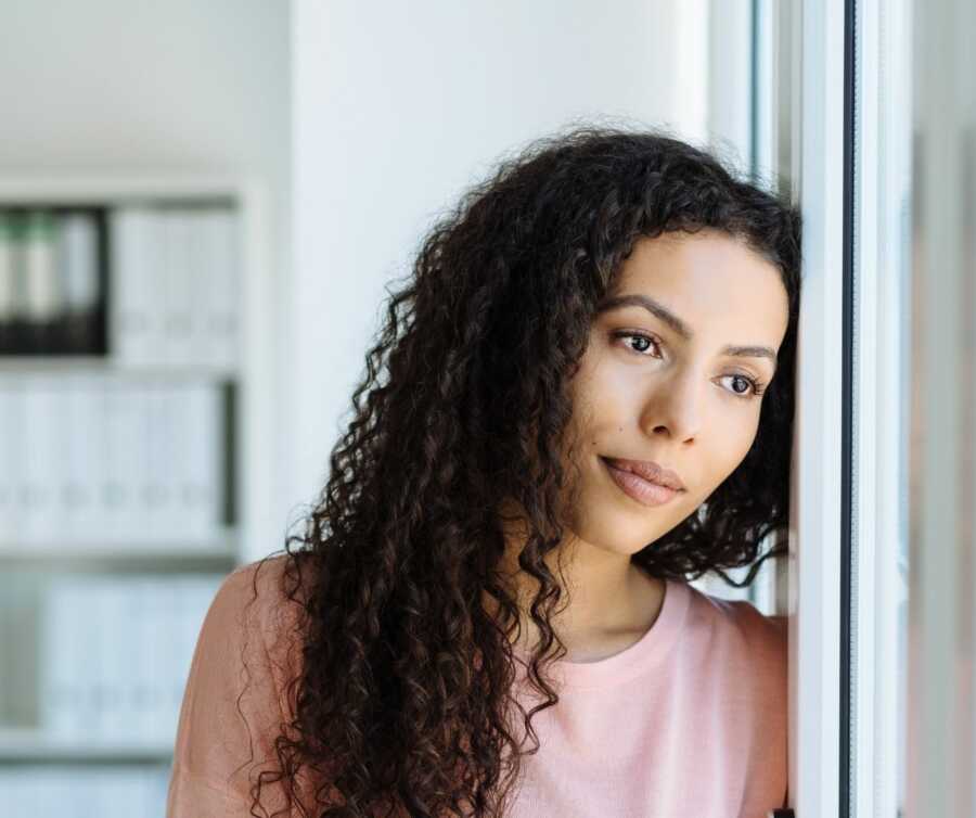 African American woman wearing pink shirt leans against the wall and stares longingly out the window.