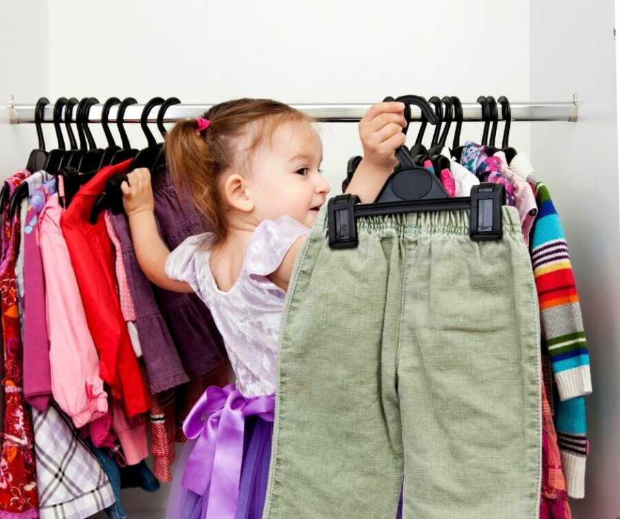 Toddler girl with pigtails wearing a purple dress holds up green pants she removed from a clothing rack in a dressing room.