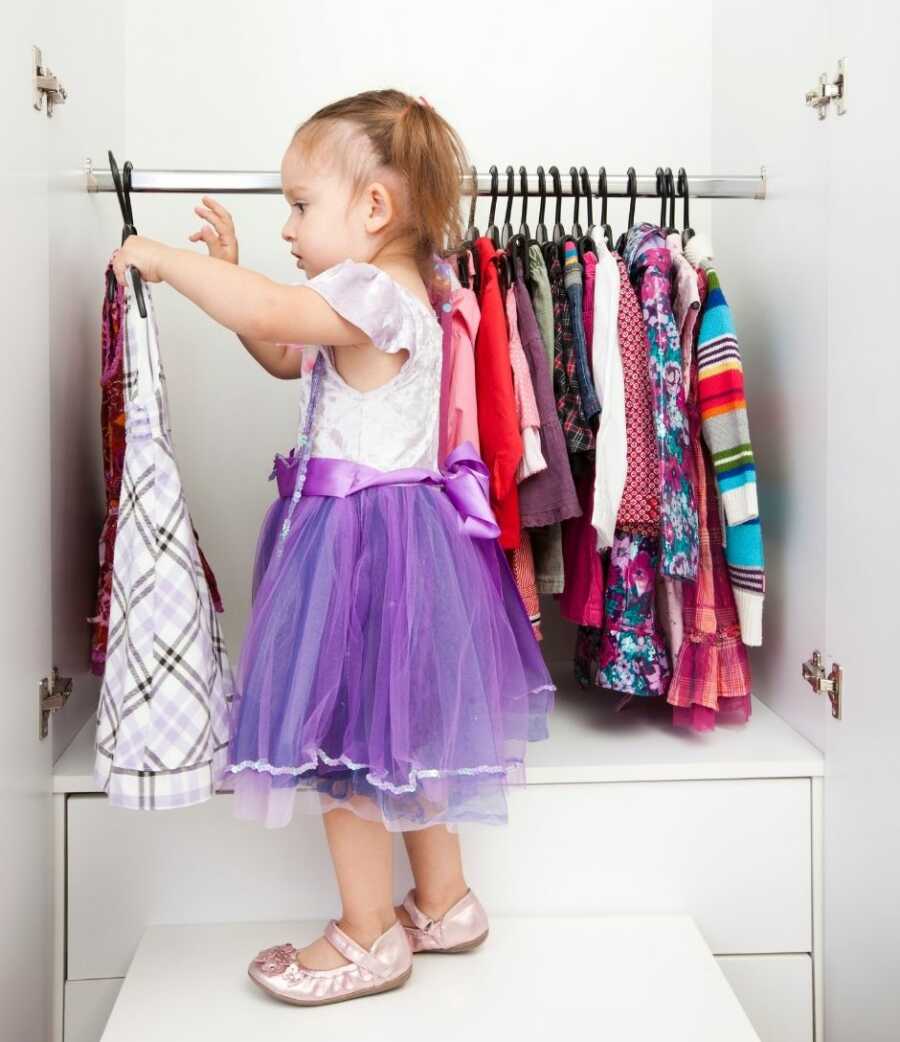 Toddler girl in purple tulle dress holds up another dress to look at in the changing room.
