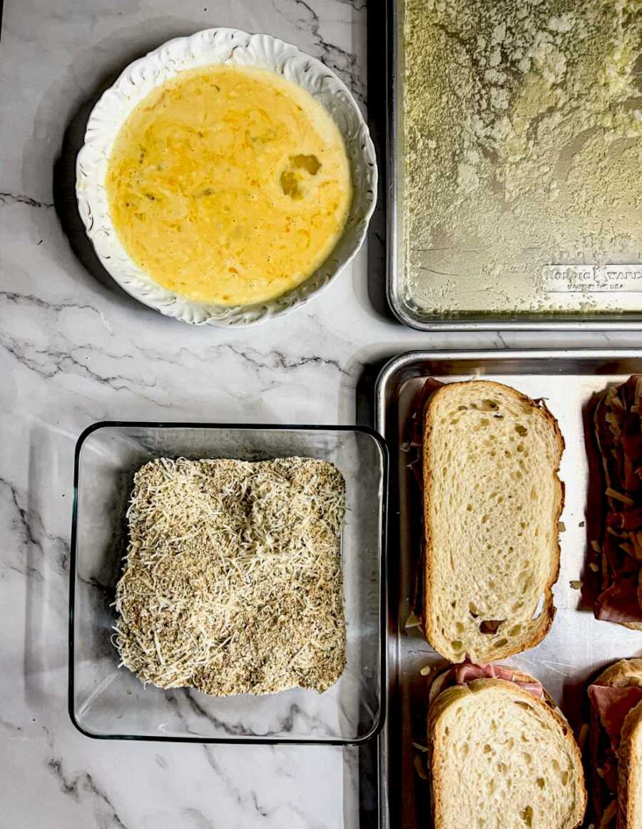 pan with sandwiches, buttered pan, egg wash bowl, and bread crumb bowl on table