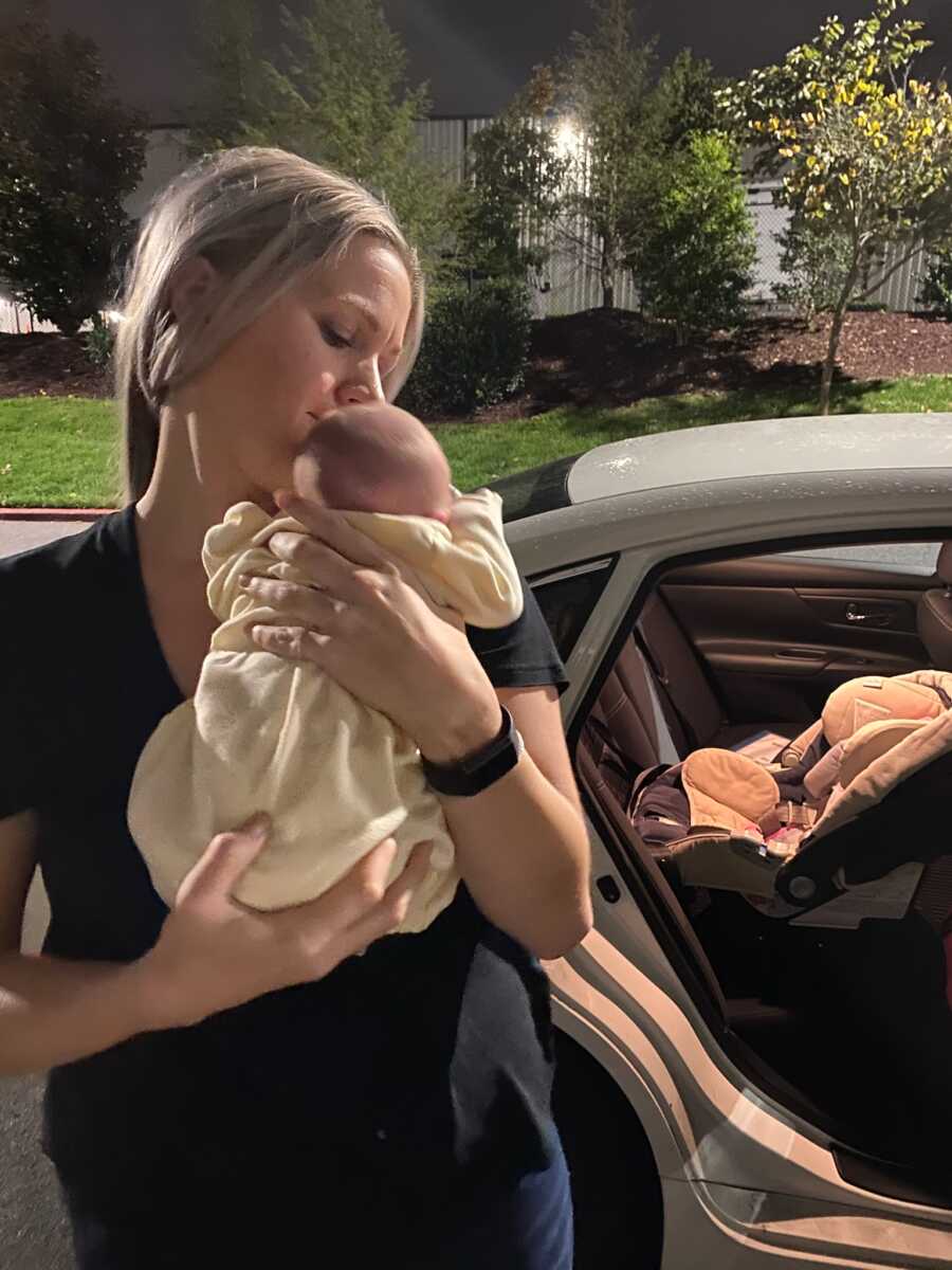 New foster mom holds infant baby girl in yellow onesie in front of social worker's car.