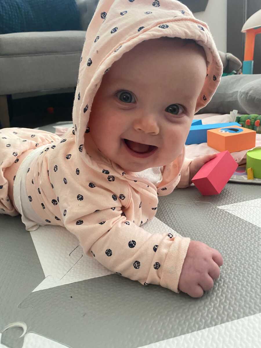 Baby foster girl lies on her tummy on the floor and smiles at the camera.