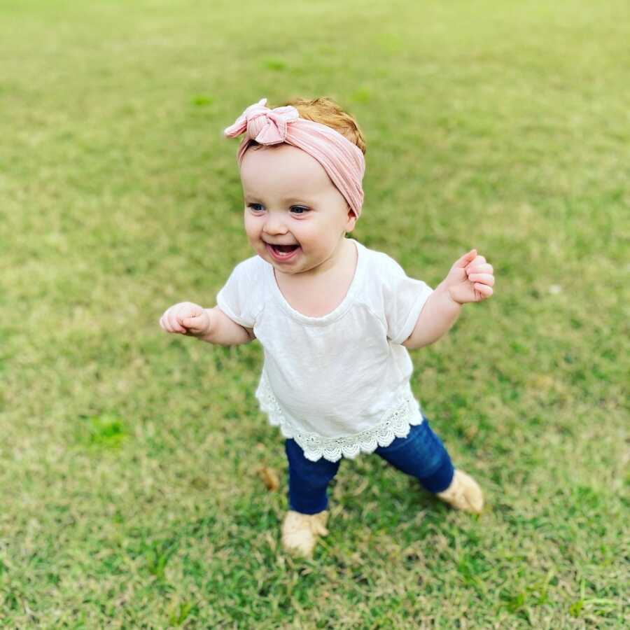 Adopted toddler girl smiles happily standing in the grass.