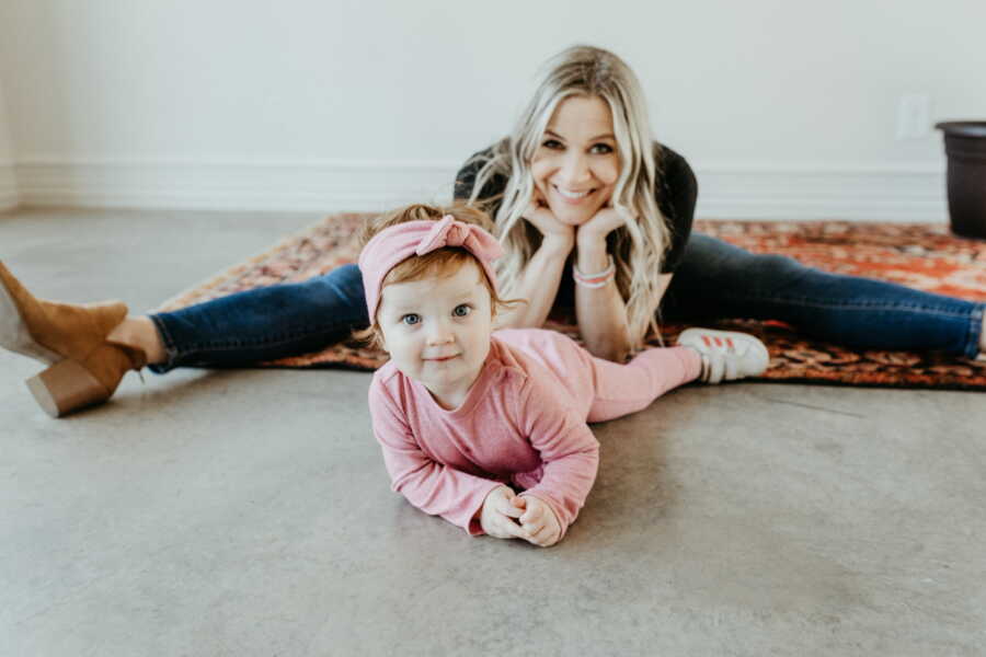 Single adoptive mom sits doing the splits and resting her chin in her hands while watching baby girl crawl on the floor in front of her.