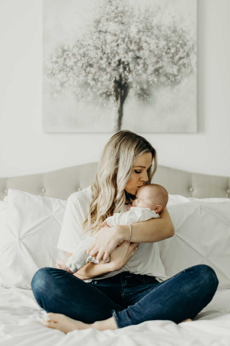 Single foster mom kisses baby girl's forehead while holding her and sitting on bed.