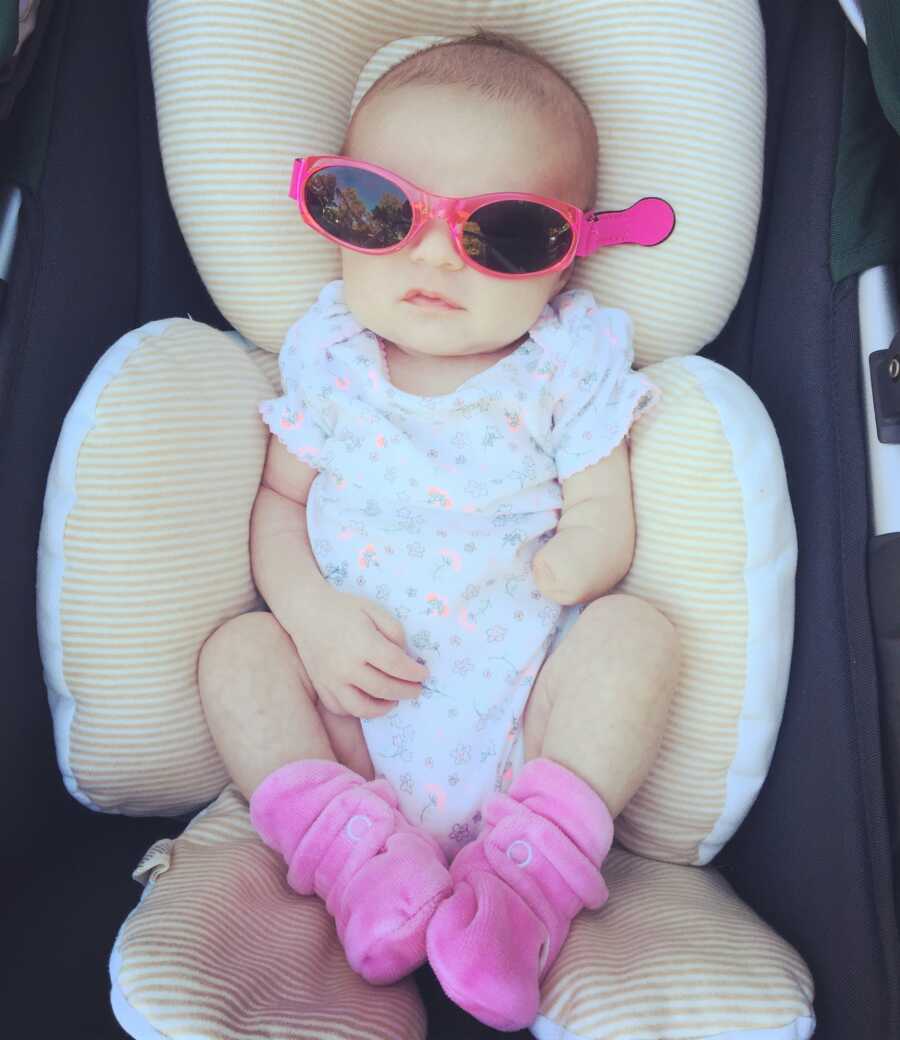 Baby girl with limb difference wears pink sunglasses and sits in car seat.