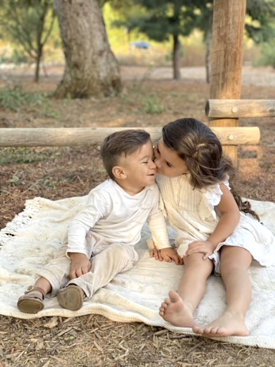 siblings sit on a blanket, sister kisses brother on the cheek