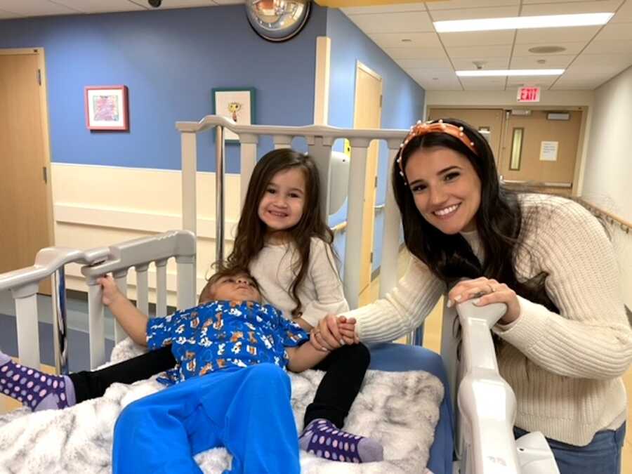 sister sits in bed with her brother while their mom stands over